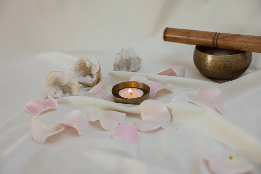 Peaceful setup with a lit candle, singing bowl, rose petals, and crystals on white linen.